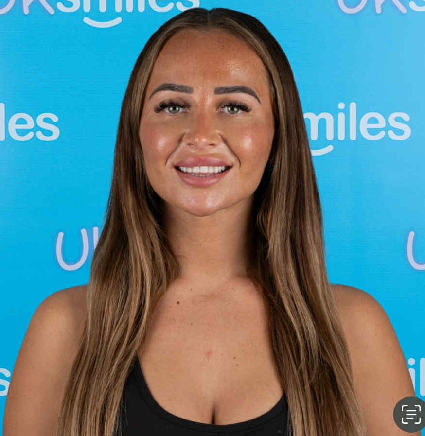 Woman with long brown hair, smiling at the camera in front of a blue background.