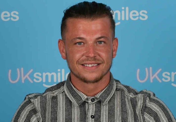 Man with short, styled brown hair smiling, wearing striped button-down shirt, against a light blue background.