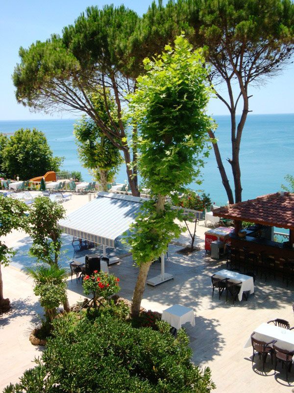 Outdoor restaurant overlooking the sea; trees, tables with white tablecloths, sunny day.