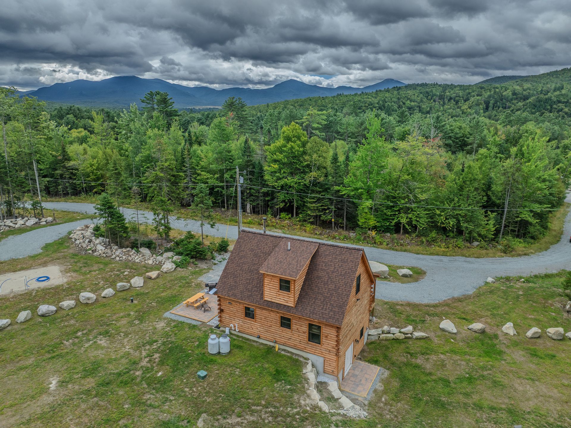 Cabin in a grassy clearing with trees, mountains in the background under a cloudy sky.