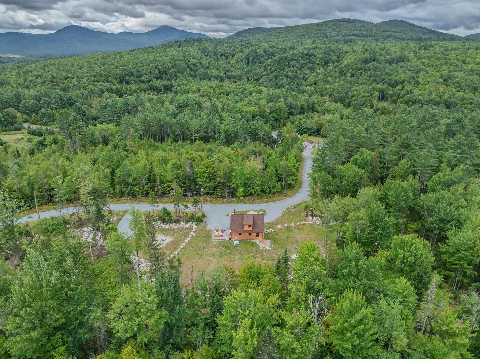 Aerial view of a small cabin and driveway surrounded by a lush green forest, with mountains in the distance.