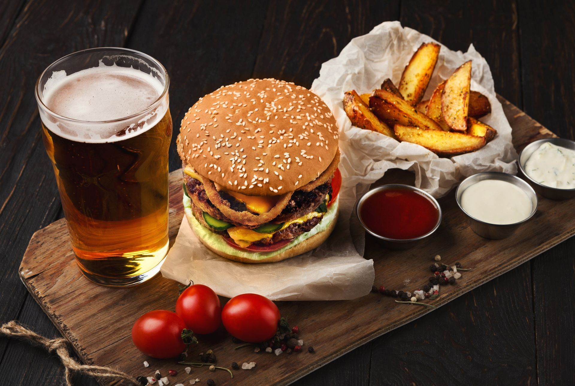 Burger, fries, beer, and dipping sauces on a wooden board with tomatoes.