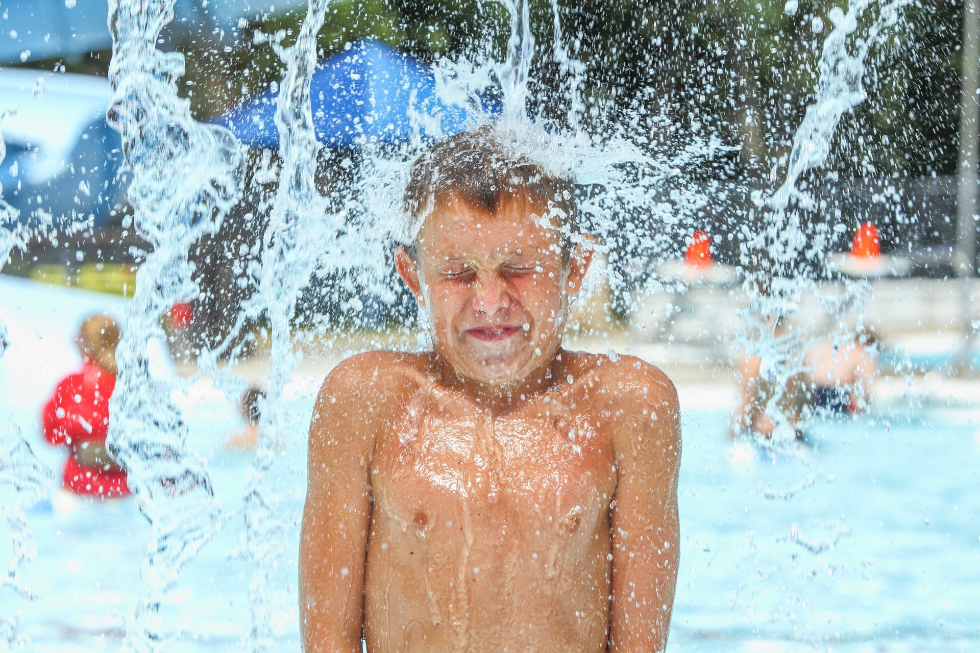 Boy getting splashed with water at a pool, eyes closed, sunny day.