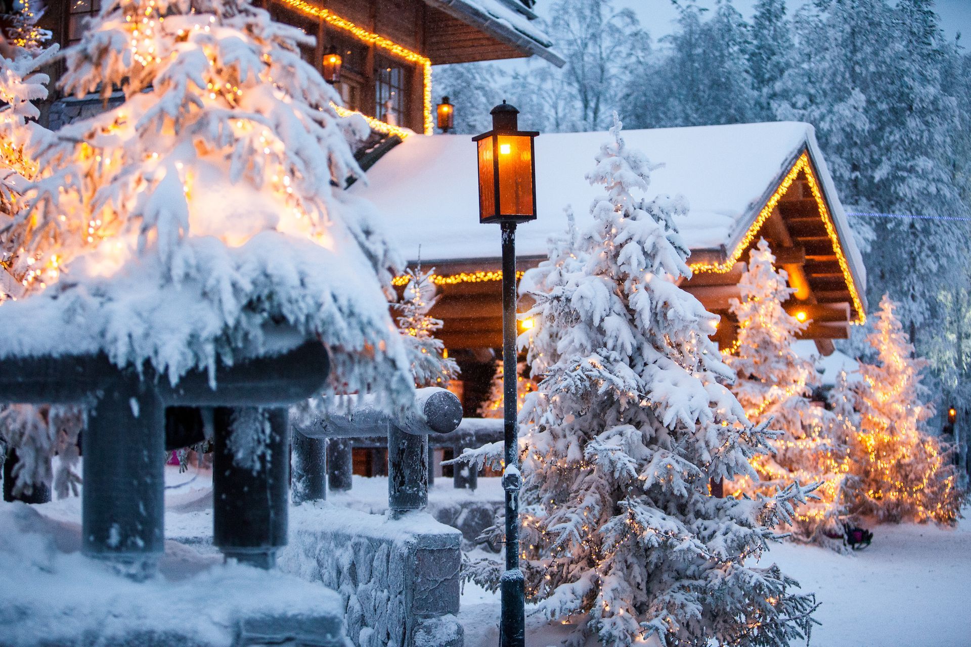 Snow-covered cabin with Christmas lights, lamp post, and snow-laden trees against a wintery backdrop.