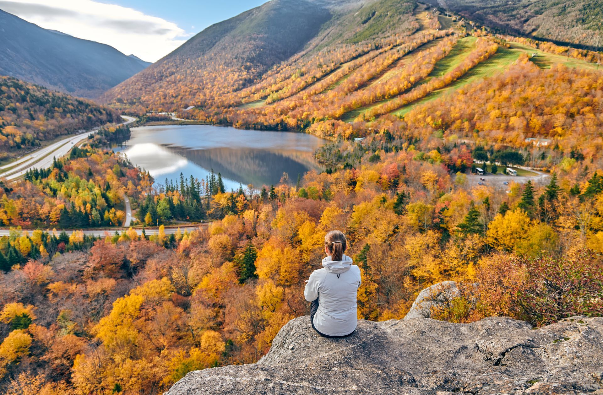 Woman overlooking a fall landscape with a lake, road, and mountains with colorful foliage.