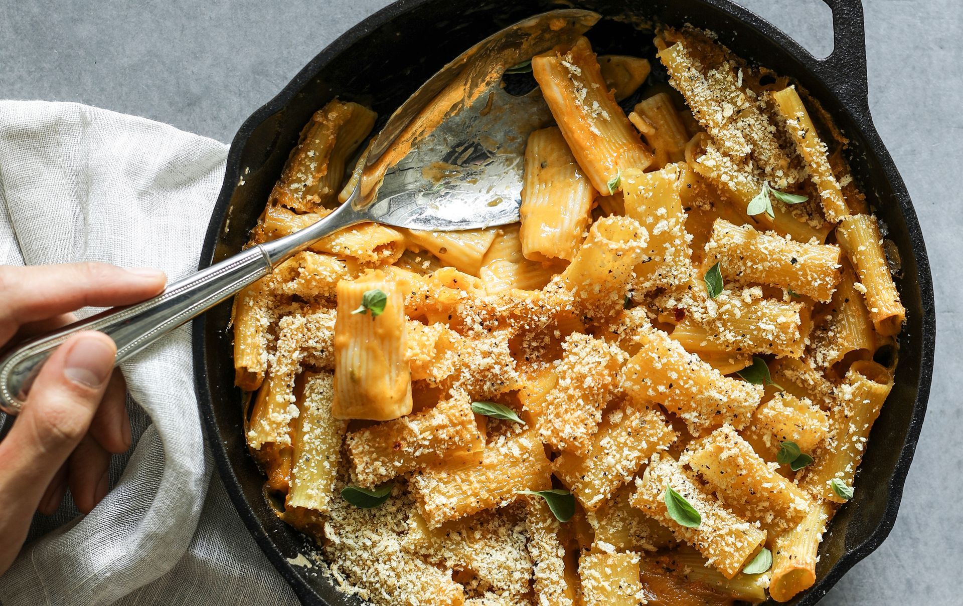 Baked pasta in a cast iron skillet with golden breadcrumb topping, being served with a spoon.