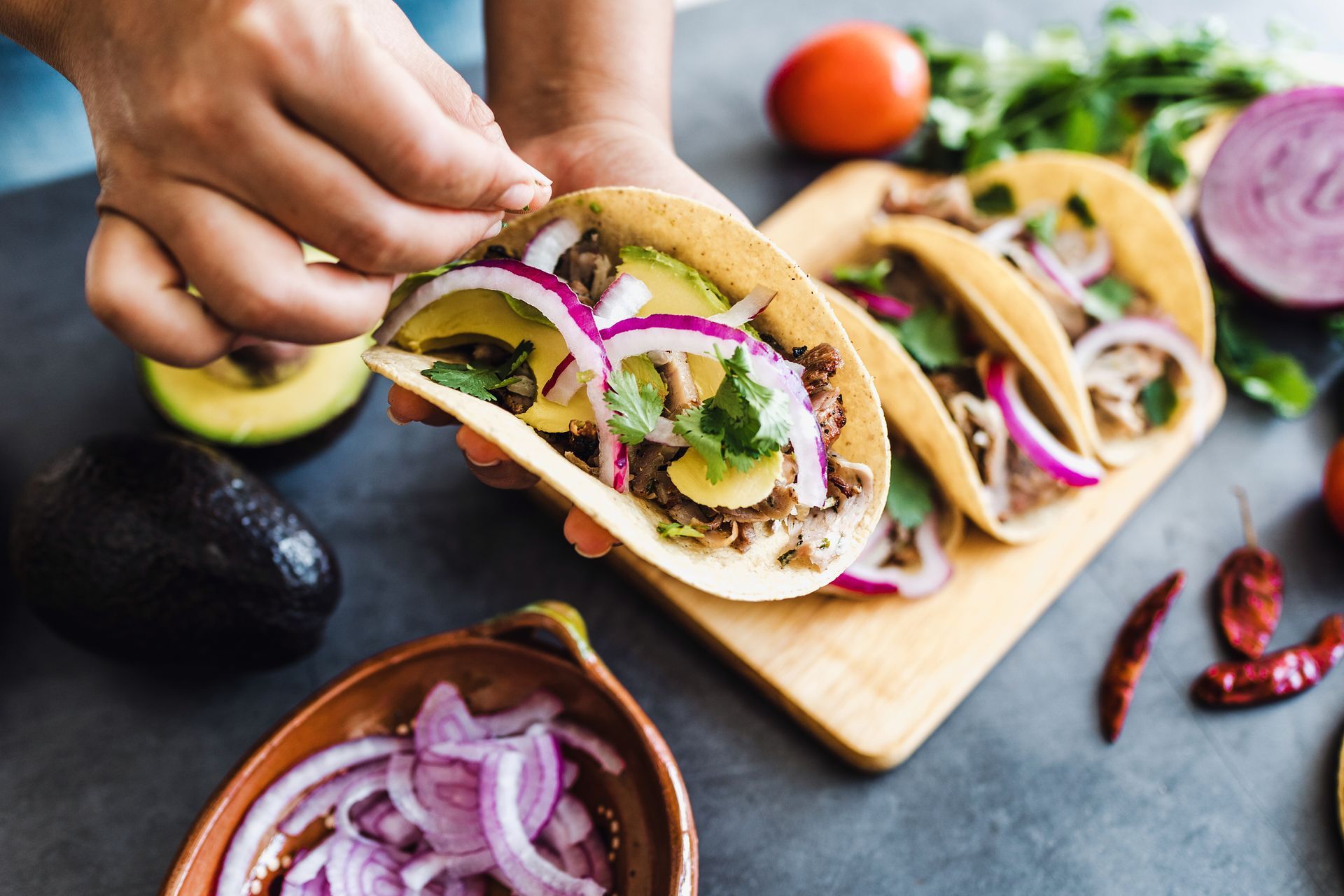 Person preparing tacos: filling tortillas with meat, avocado, red onion, and cilantro.