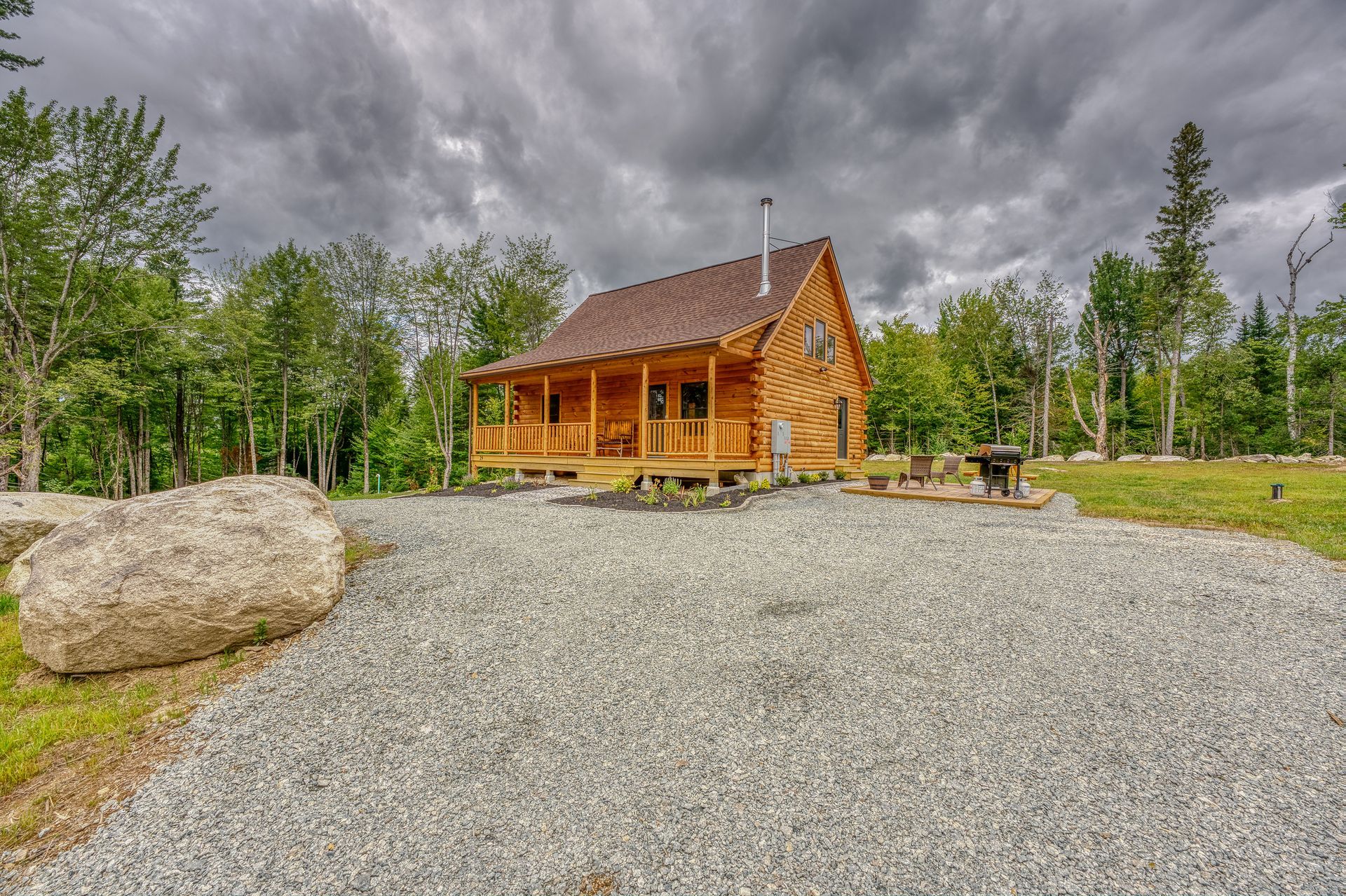 Wooden cabin with porch, set in a gravel yard under a stormy, overcast sky.