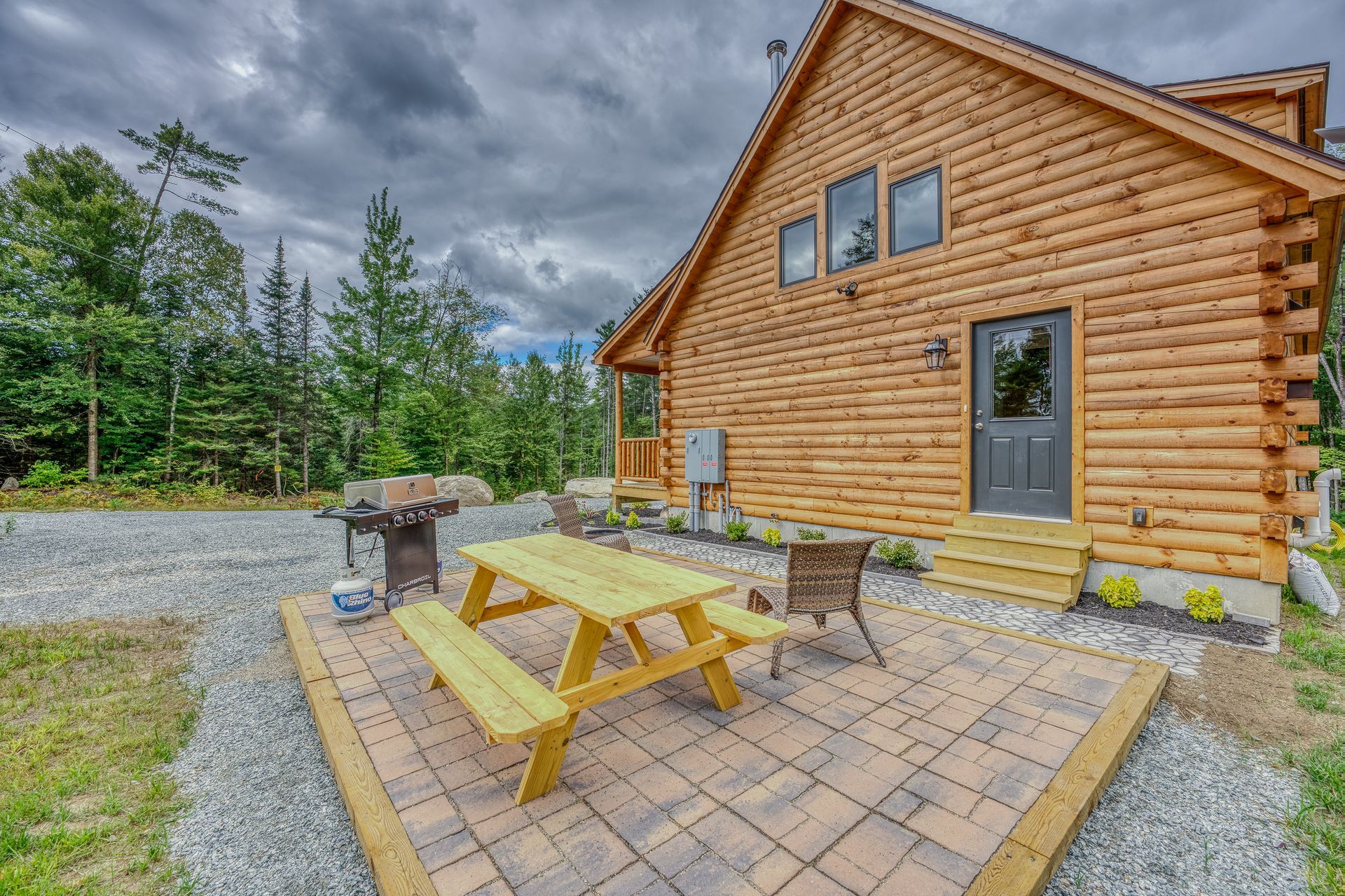 Log cabin exterior with patio, picnic table, and grill, surrounded by trees. Overcast sky.