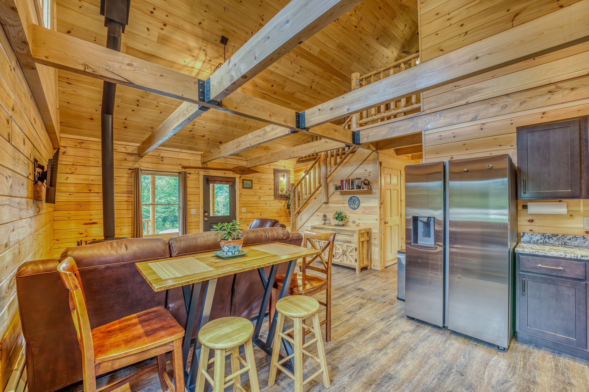 Interior of a log cabin with a kitchen, dining area, and living room, with exposed wood beams and a stainless steel refrigerator.