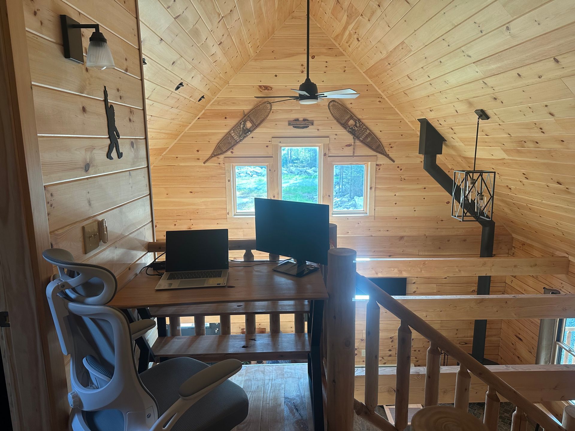 Cozy wooden loft workspace with monitors, chair, snowshoes, and windows.