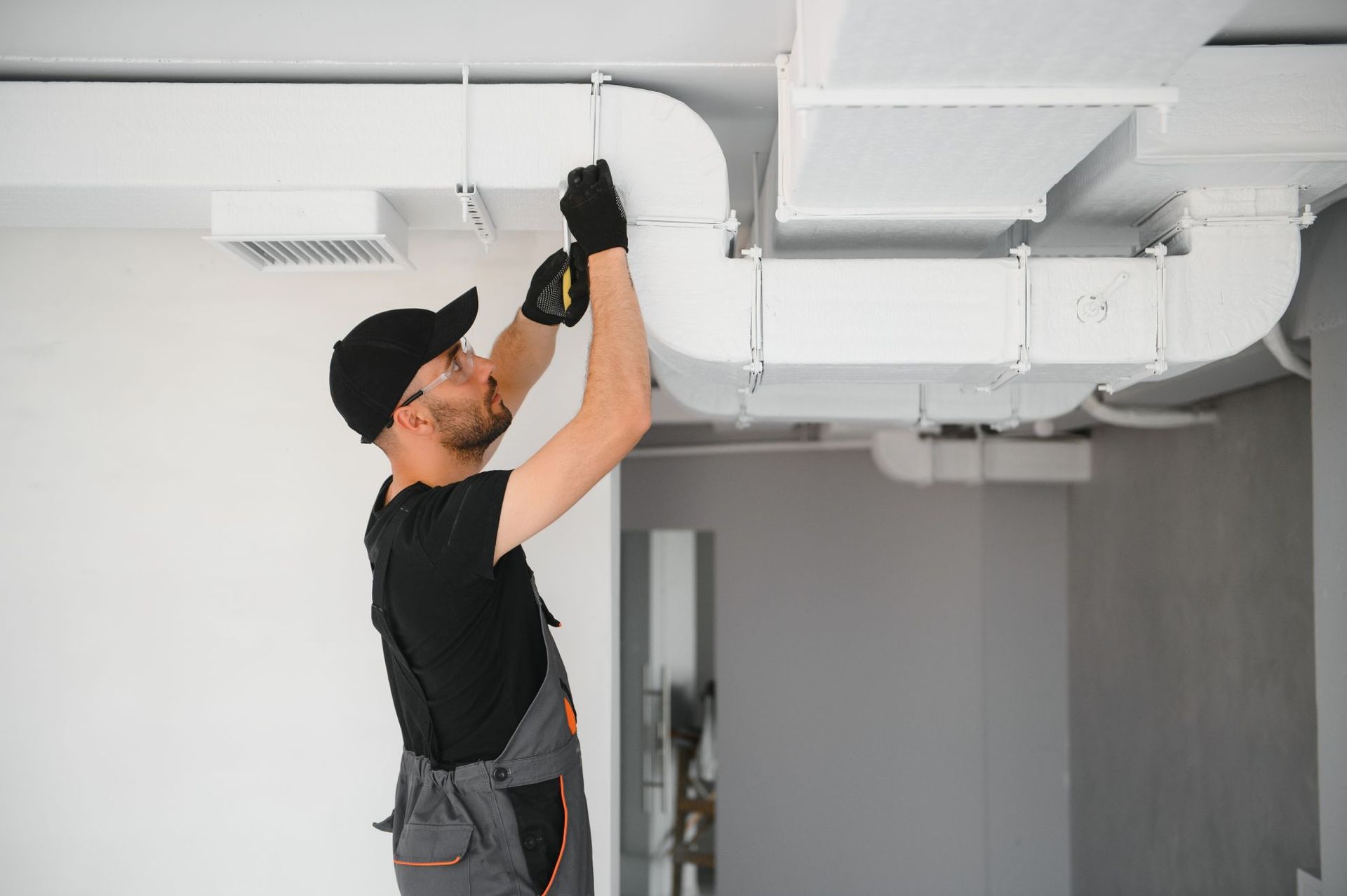 Worker inspecting white ceiling ductwork in a modern room, wearing a black cap and gloves.