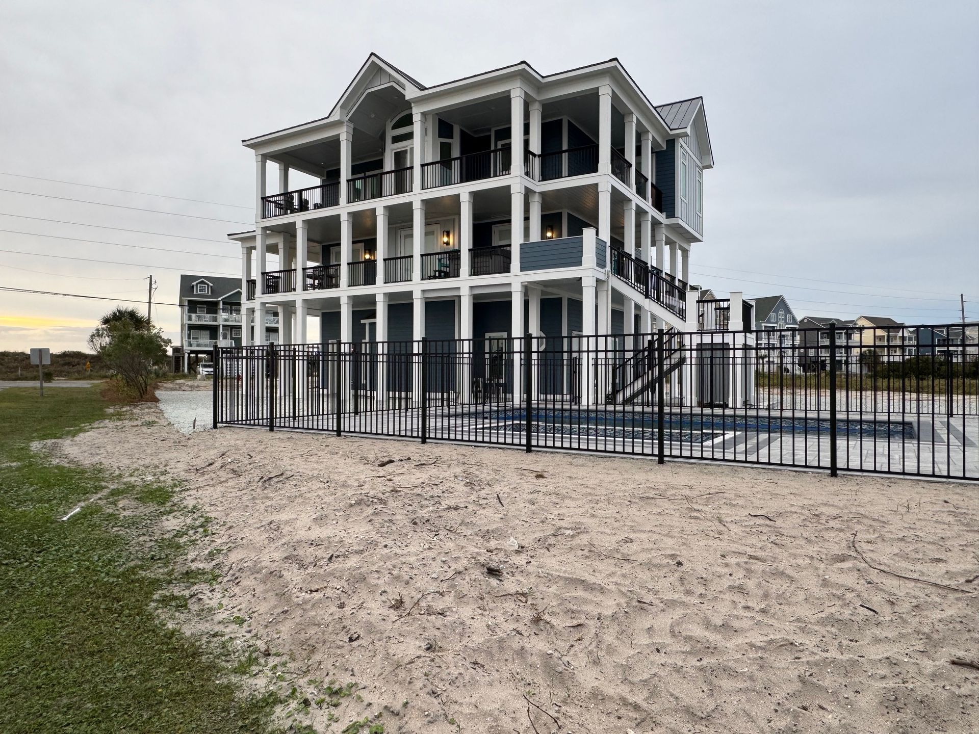 white with black trim, on stilts with a black fence, and a sandy yard.