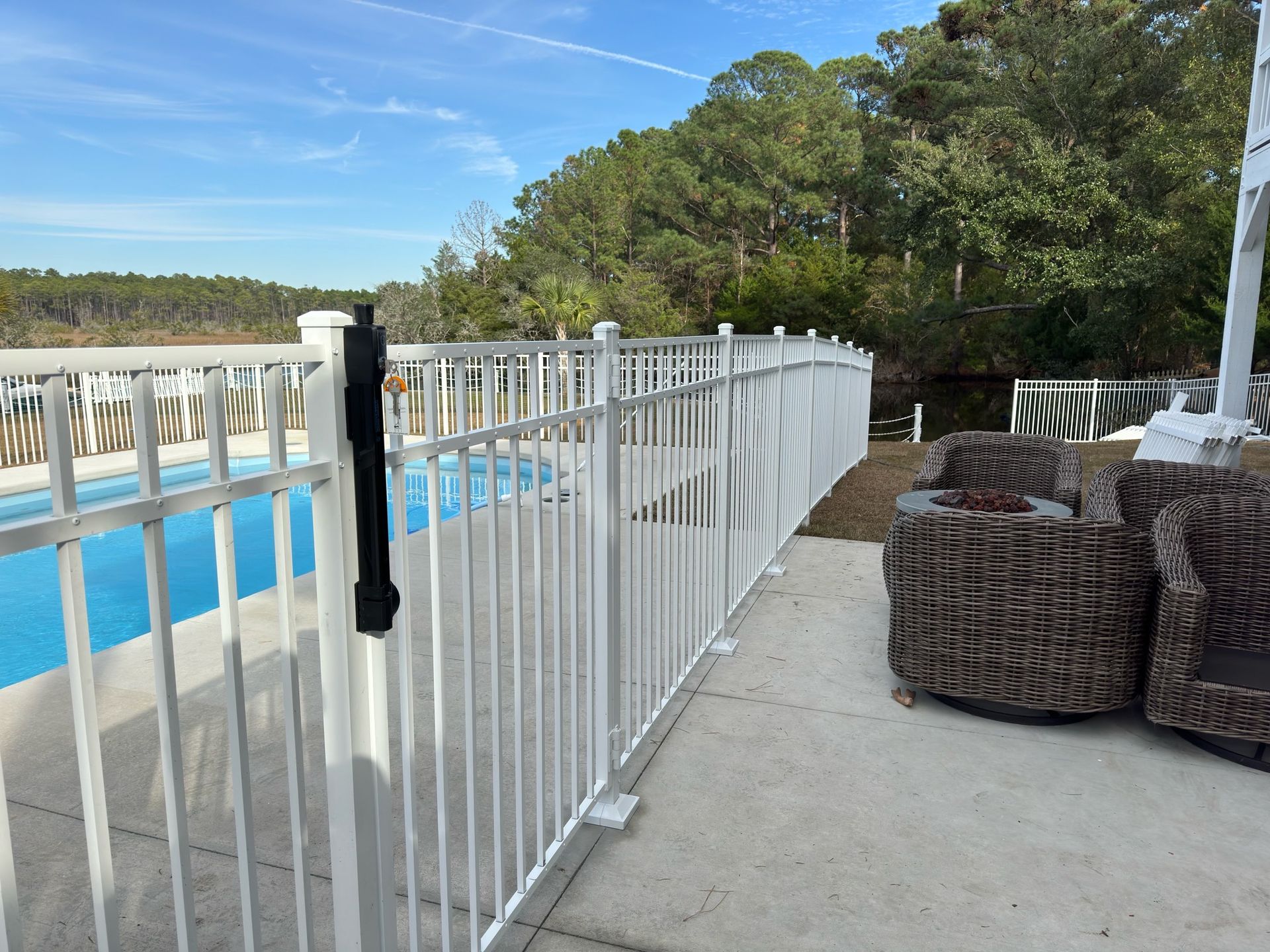 White fence surrounds a blue pool with trees and chairs in the background on a sunny day.