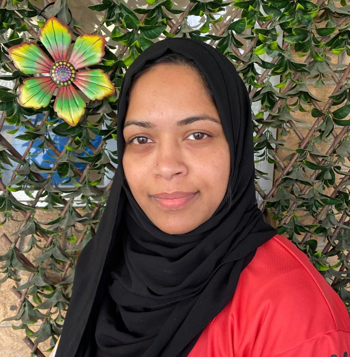 A woman wearing a black scarf and a red shirt is standing in front of a flowered wall, she the class leader.