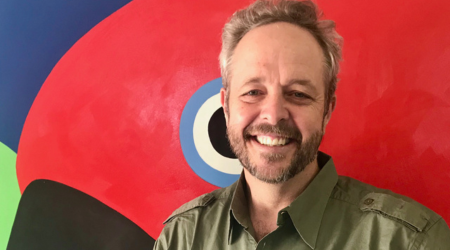 Man smiles, posing in front of a colorful, abstract bird mural. He wears a green button-up shirt.
