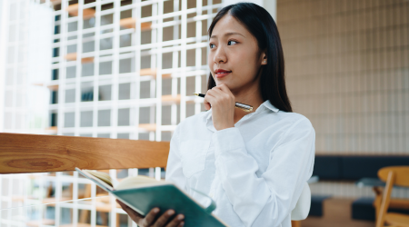 Woman in white shirt thoughtfully holding a pen and notebook, looking away from window.