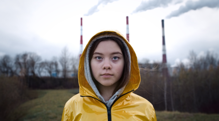 Person in yellow raincoat, facing forward, in front of smokestacks, cloudy sky.