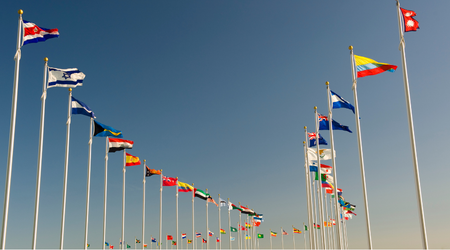 Flags of various nations on tall poles, waving in the wind under a clear blue sky.