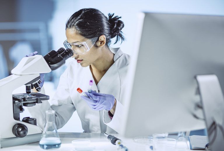 Scientist in lab coat looks into microscope, with gloves and computer, examining a sample.