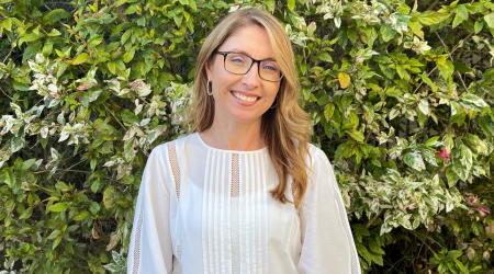 Woman with glasses smiles in front of green and white foliage, wearing a white blouse.