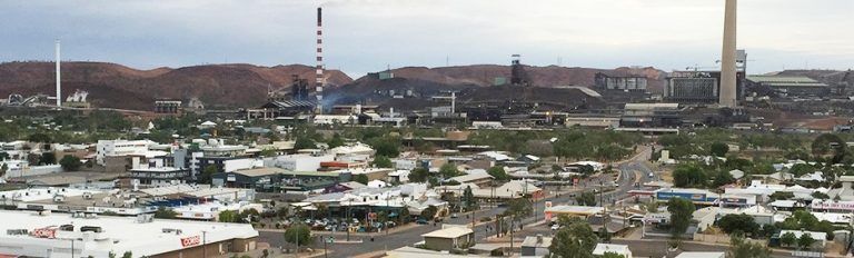 A cityscape with industrial structures, buildings, and a mountain range under a cloudy sky.
