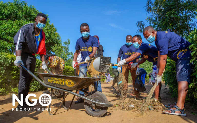 People in blue shirts and masks cleaning up with a wheelbarrow and brooms outdoors.
