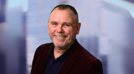 Man with short hair smiles at the camera, wearing a dark blue shirt and maroon jacket, in front of a blurry background.