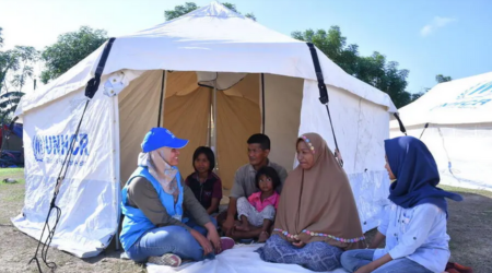 Group sits in tent; UNHCR worker speaks. Outdoors, light-colored tent.