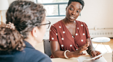 Woman in a polka dot top gestures while talking to another woman at a table.
