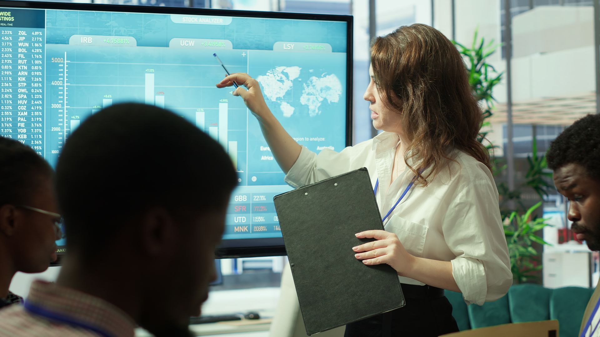 Woman presenting charts and world map on screen, pointing with pen in a meeting.
