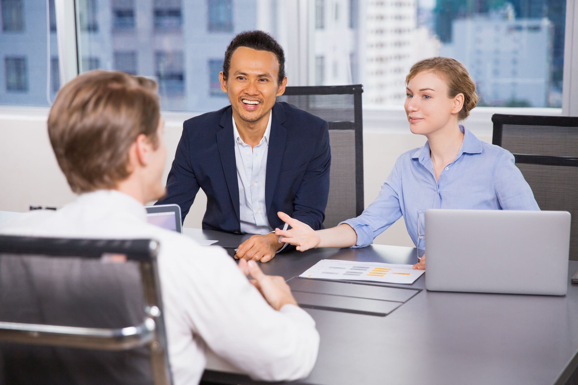 Three people in a meeting. A man and a woman face another man at a table, gesturing, with a laptop and papers.