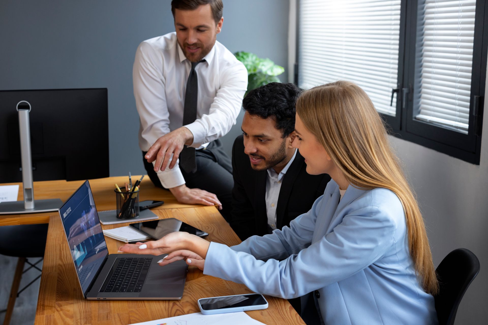 Three people collaborate around a laptop at a desk; a woman points, men look on.