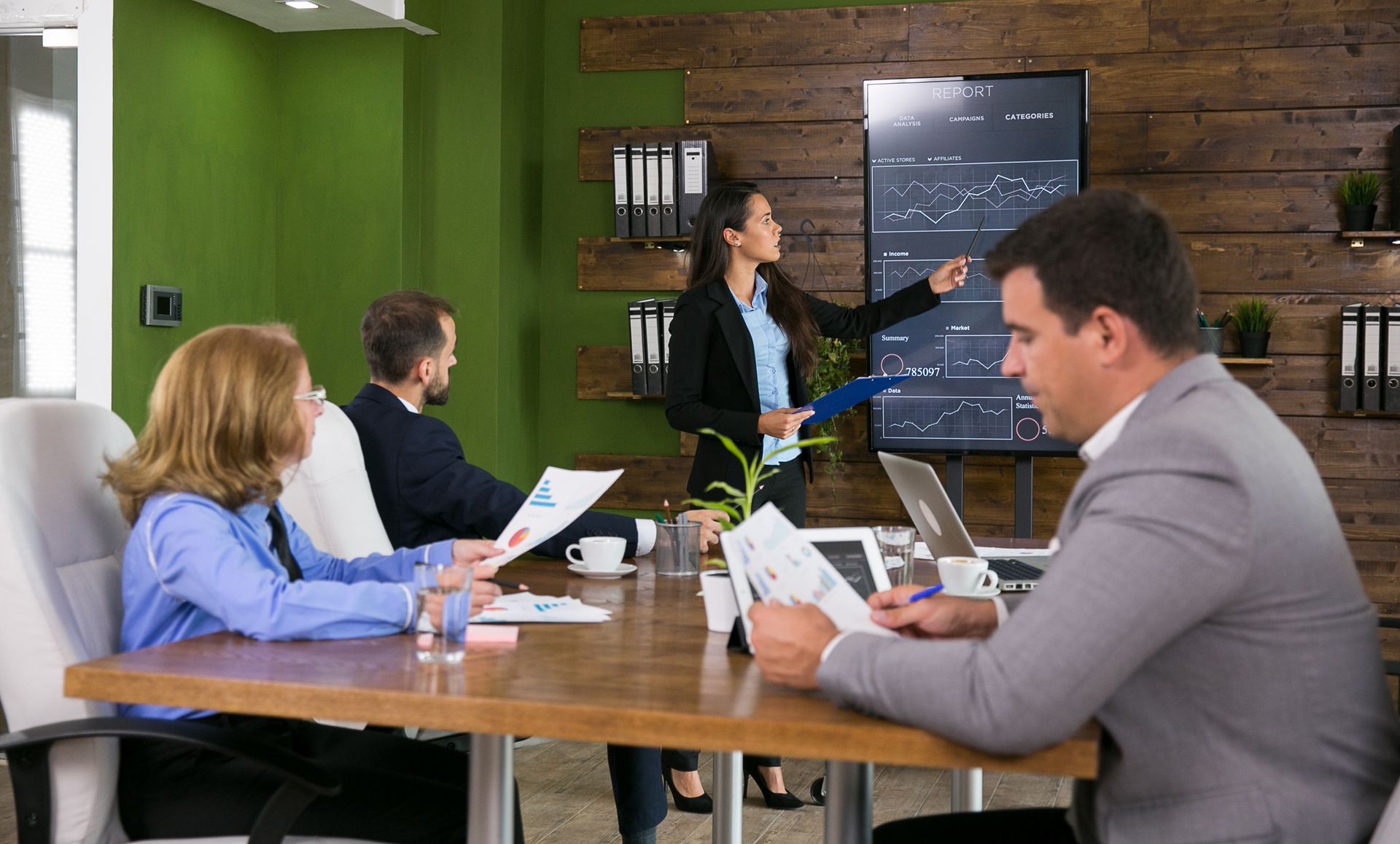 Business team in a meeting, woman pointing at graph on screen, reviewing documents at table.