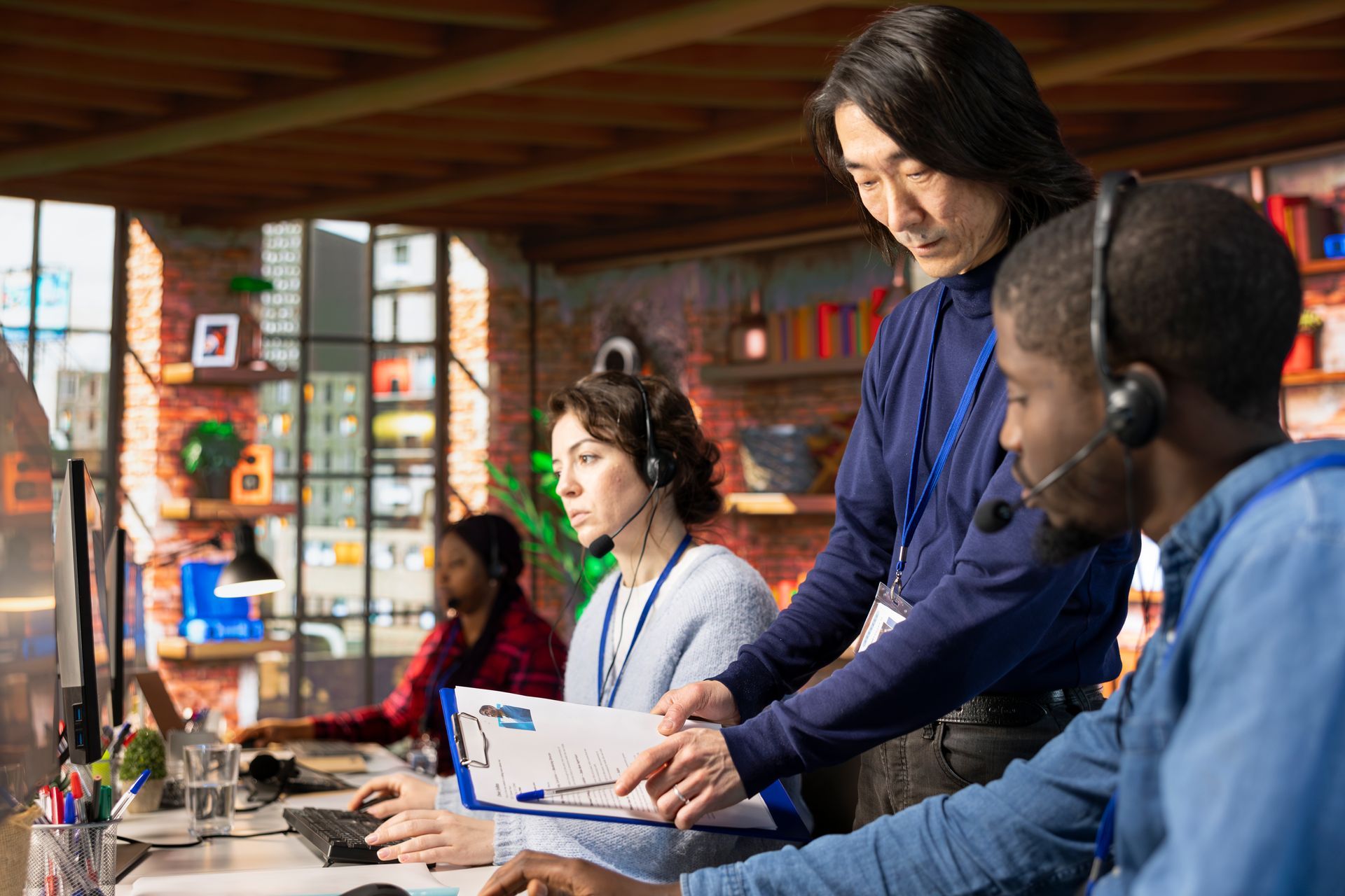 Colleagues working at computers with headsets. One person assists another while holding a clipboard. Interior shot.