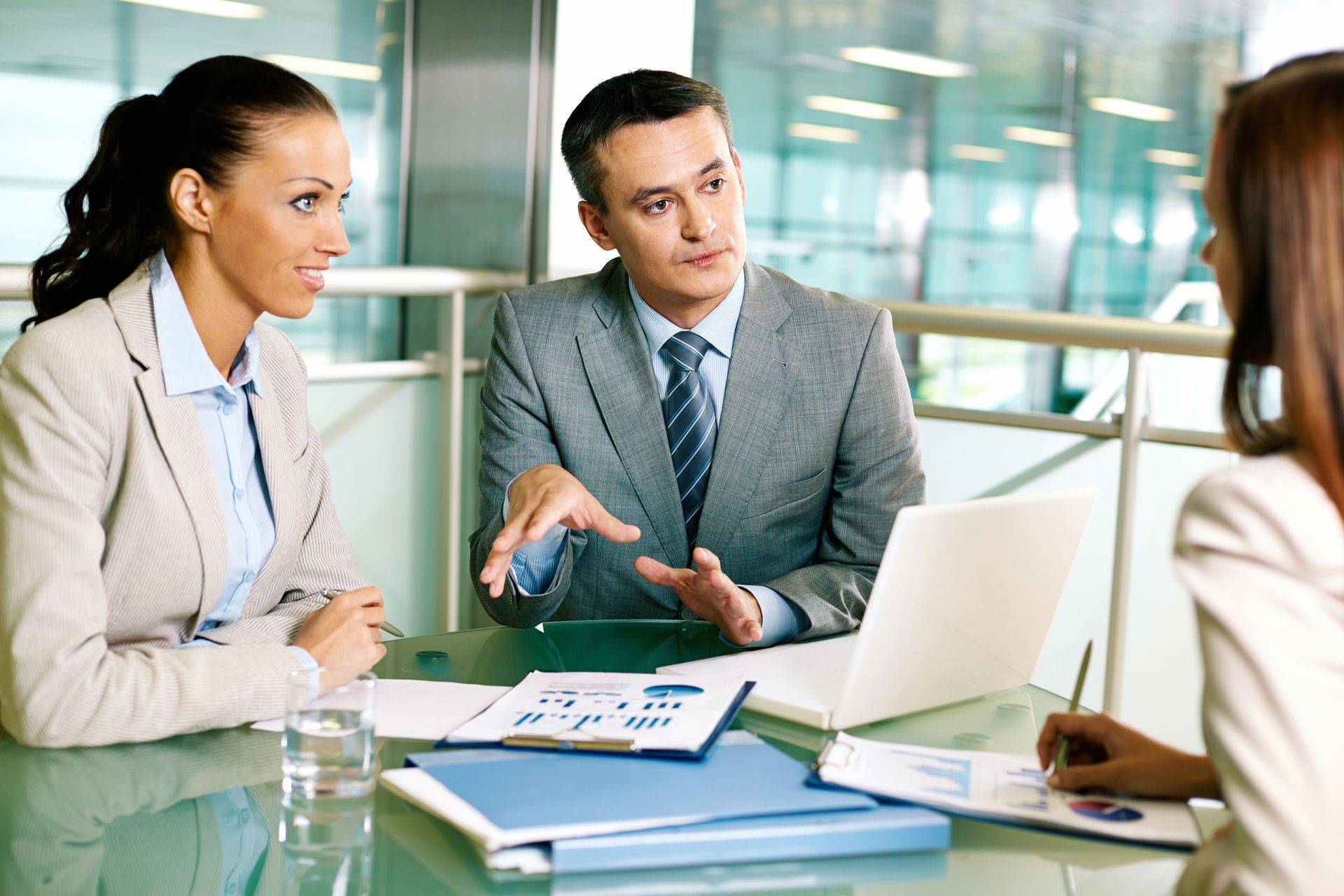 Three professionals in business attire at a table, discussing documents and a laptop in a modern office.