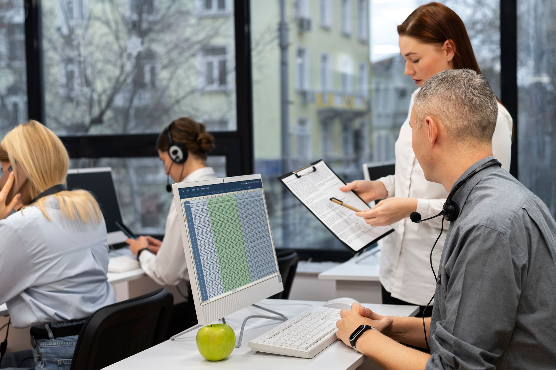 Office setting with employees wearing headsets, one being assisted by a colleague with a clipboard near a computer.