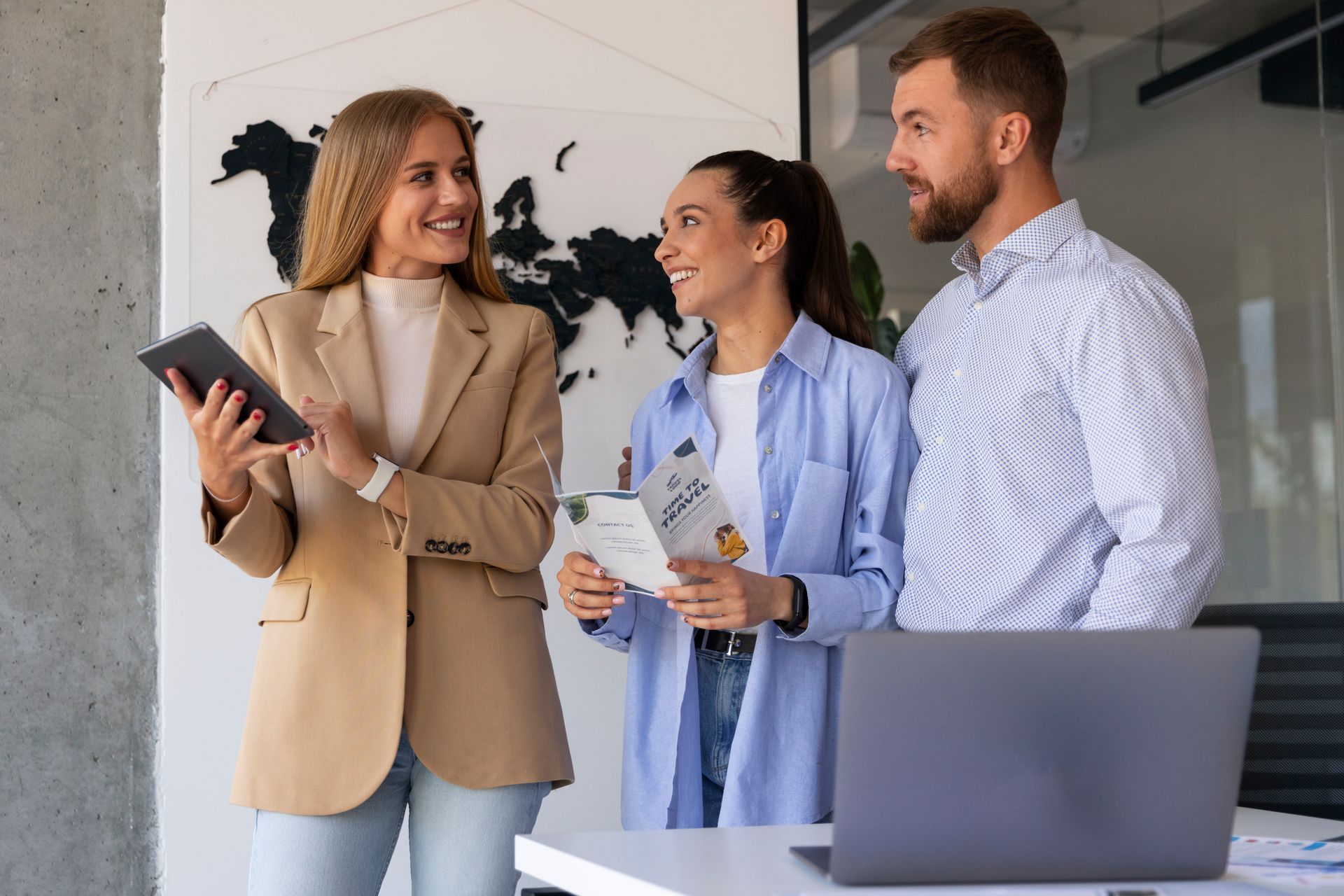Three coworkers in an office setting discussing documents and tablet. Woman in tan blazer smiles. World map in background.