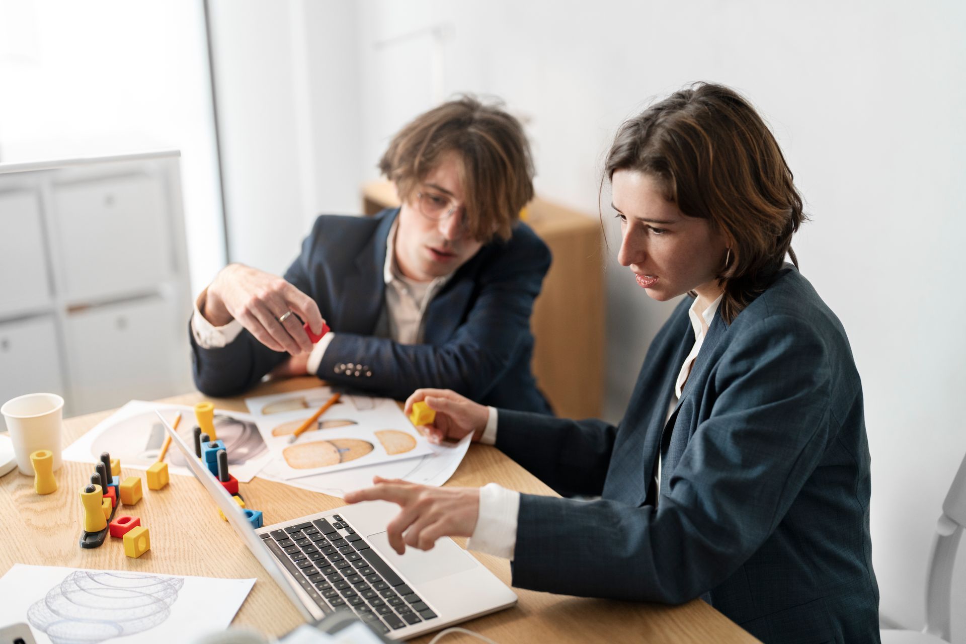 Two people in suits looking at a laptop and documents on a wooden desk.
