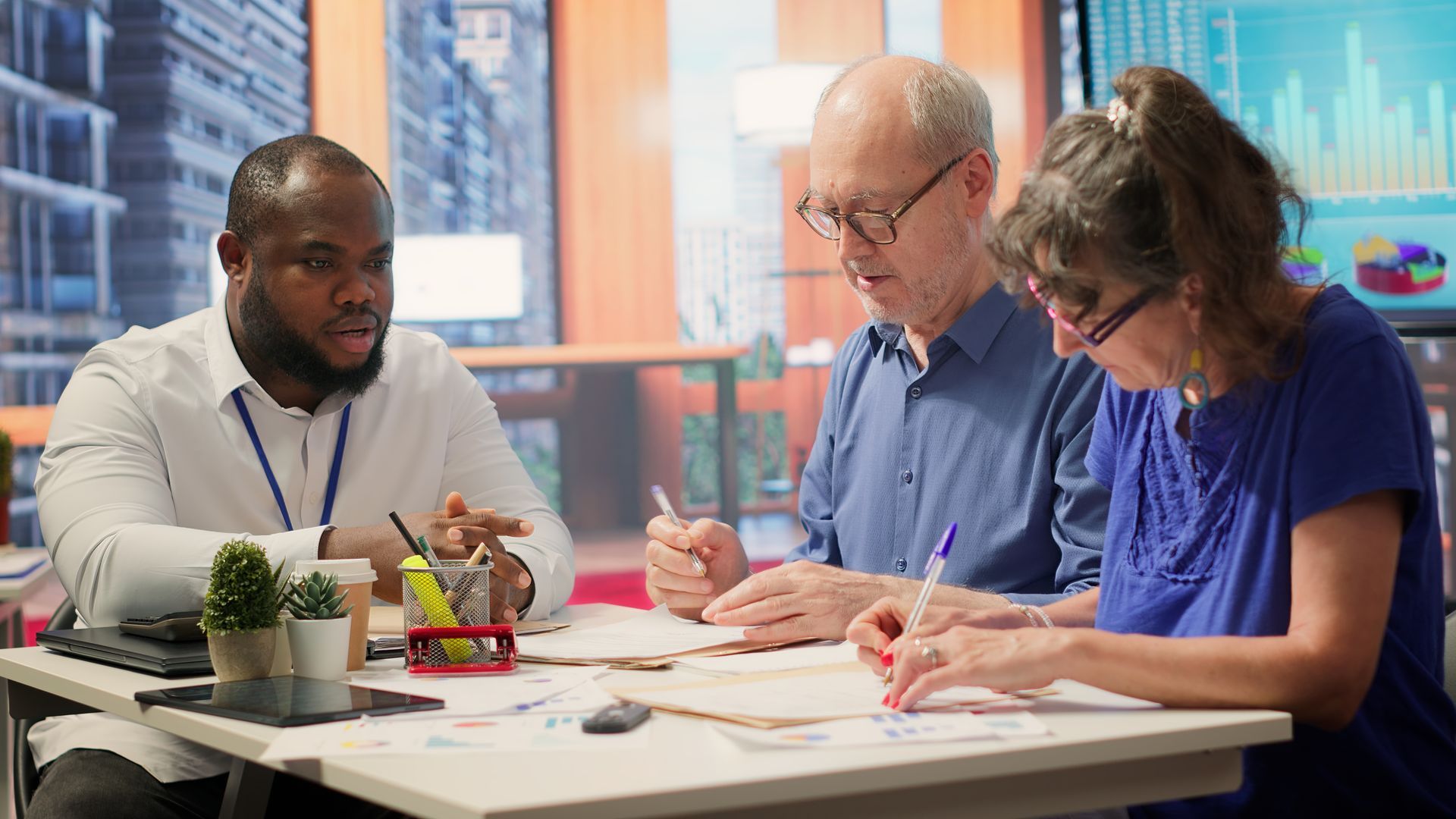 Three people review documents at a table; one speaks while others write; charts on a wall in background.