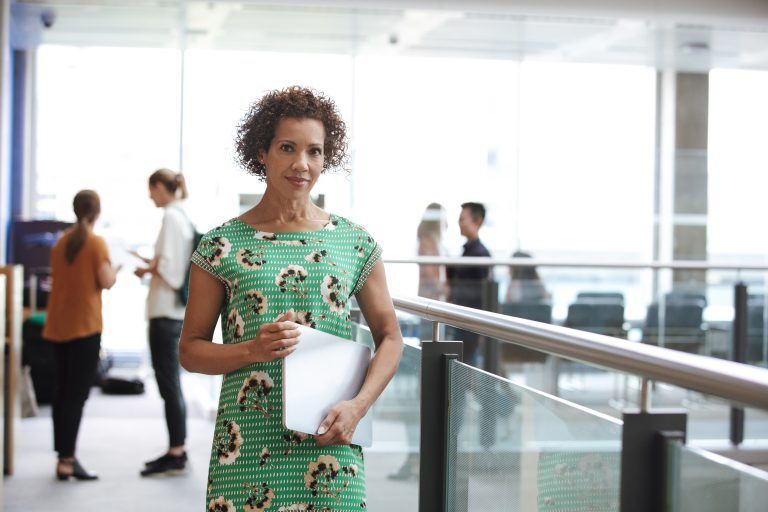 Woman in green dress holds papers, standing in a brightly lit hallway; two people in the background.
