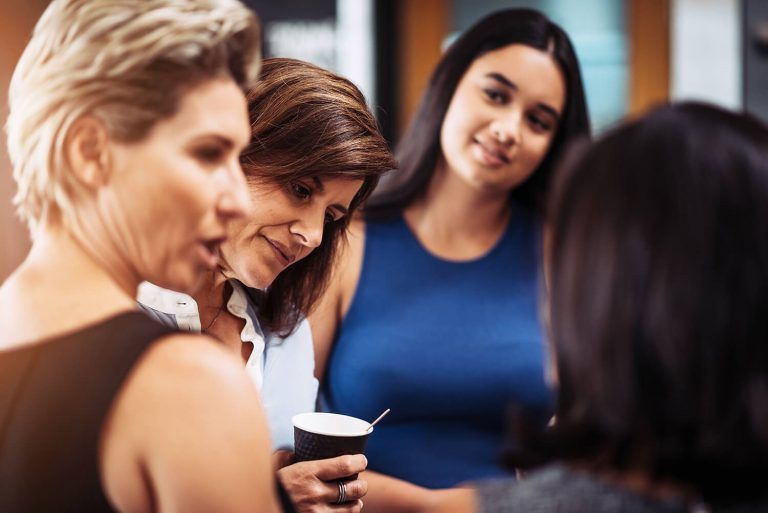 Four women in discussion, two are looking at each other, one holds a coffee cup, and another is in the background smiling.