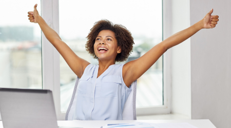 Woman at desk with arms raised, giving thumbs up. Smiling, excited expression.