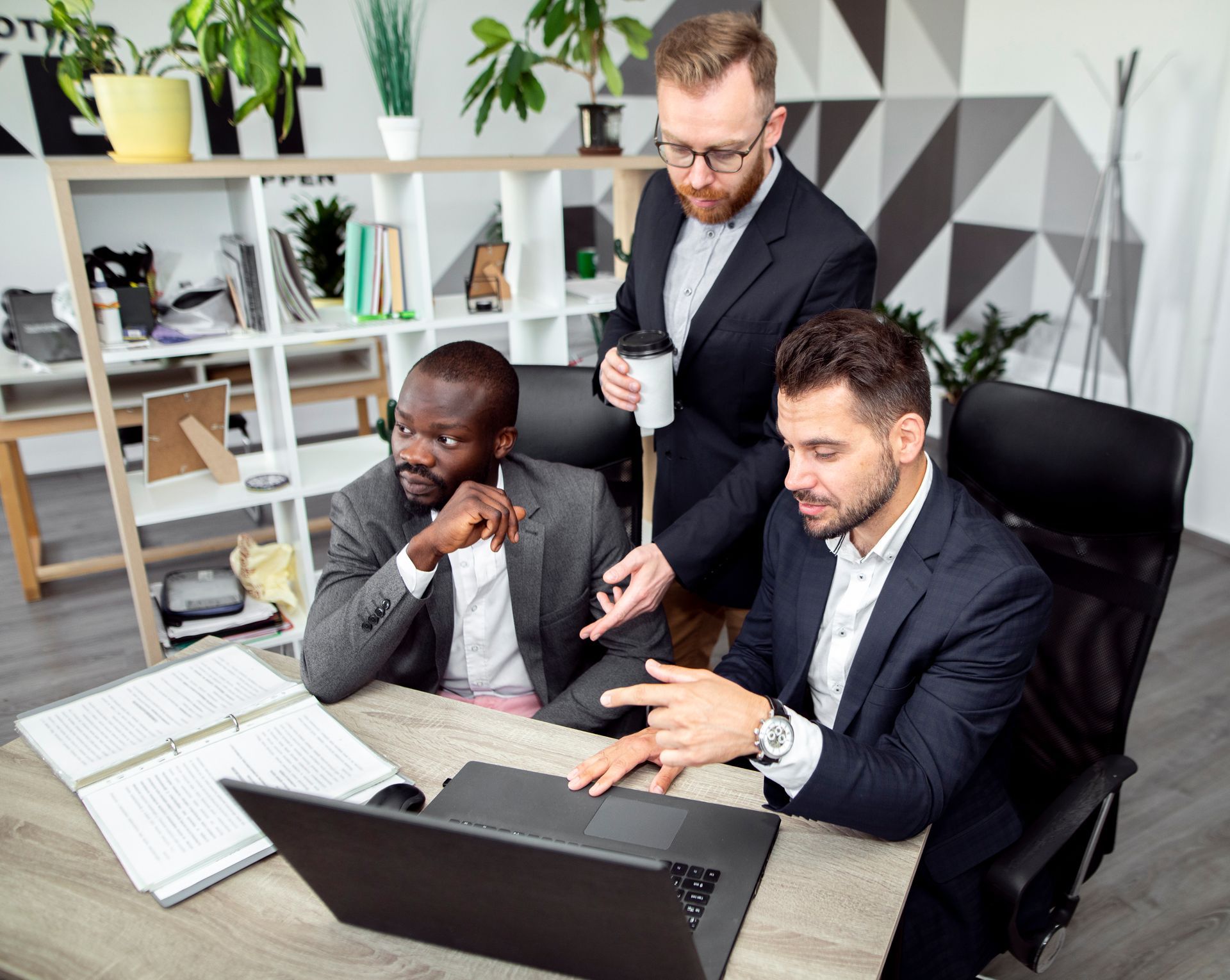 Three businesspeople in suits looking at a laptop at a desk in an office. One points at the screen.