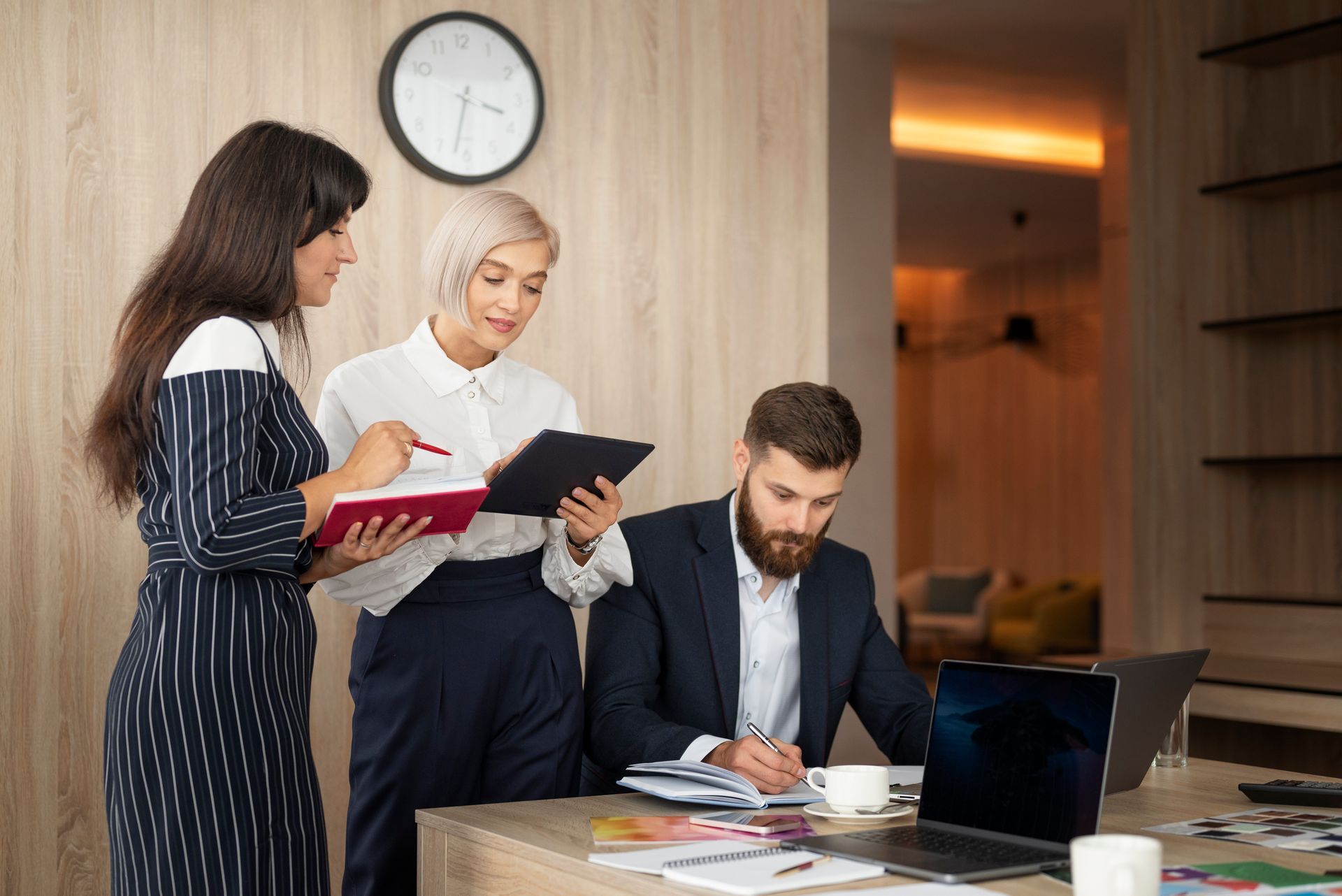 Three people collaborating in an office. One holds a red notebook, another a tablet; a man writes at a desk.