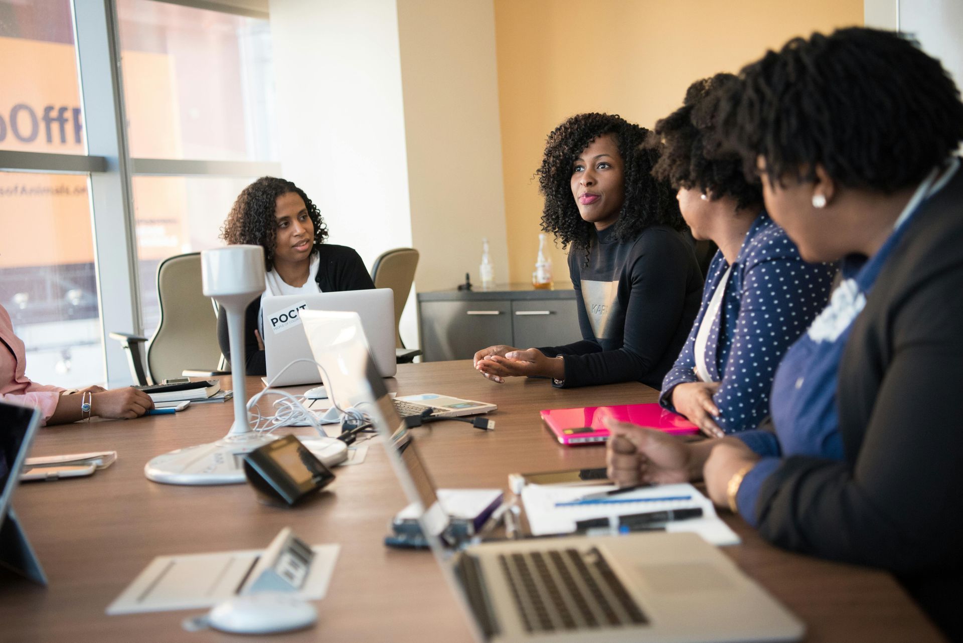 Women in a business meeting, gathered around a conference table with laptops and papers.