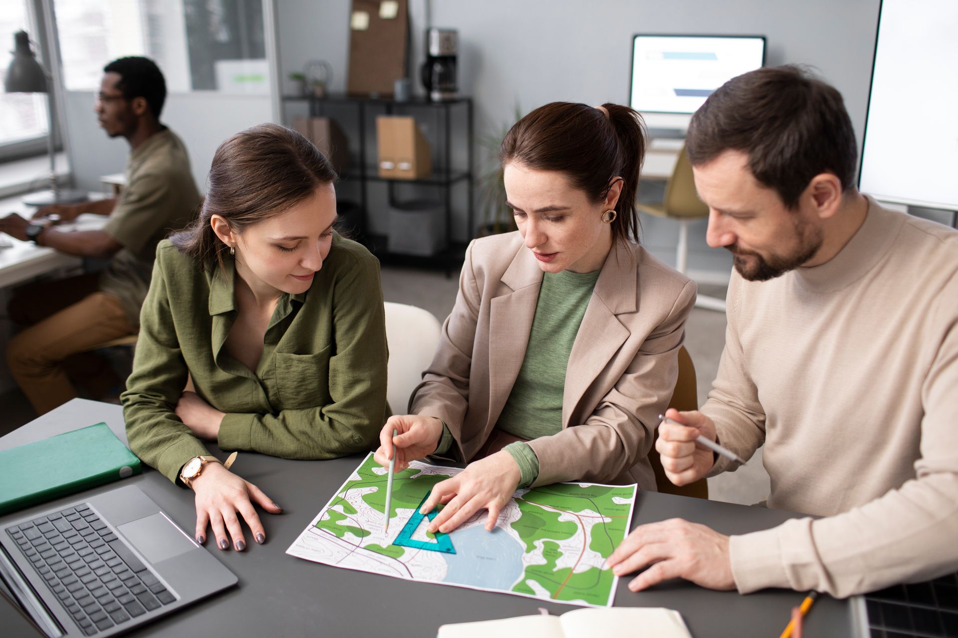 Three colleagues examining a map at a desk, pointing and discussing. Another person works at a computer in the background.