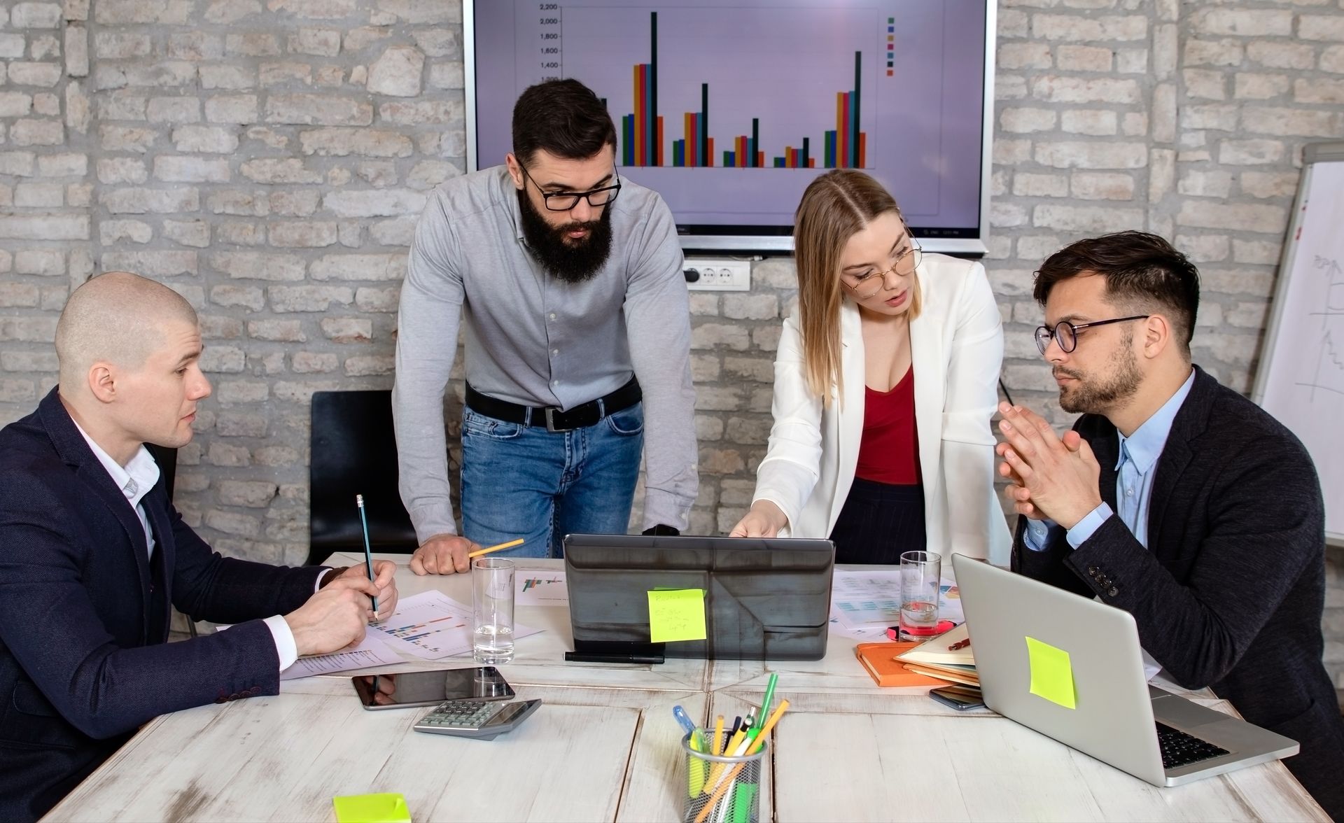 Four businesspeople in a meeting, examining data on a table, with a graph displayed on a screen in the background.