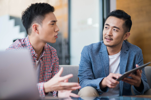 Two men in discussion, one gesturing and the other holding a tablet in a modern office setting.