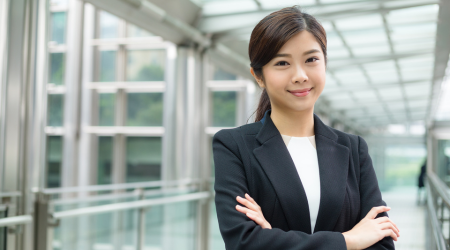 Woman in business suit, arms crossed, smiling in an office hallway.